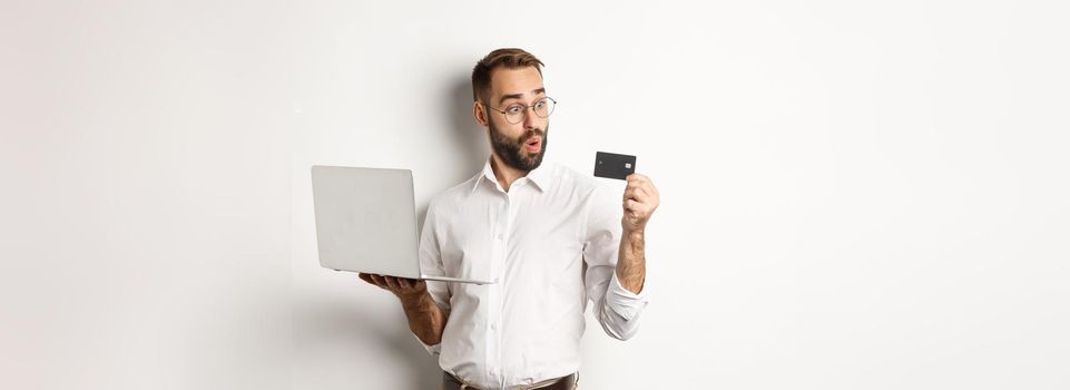 Online shopping. Amazed businessman holding laptop, looking impressed at credit card, standing over white background.