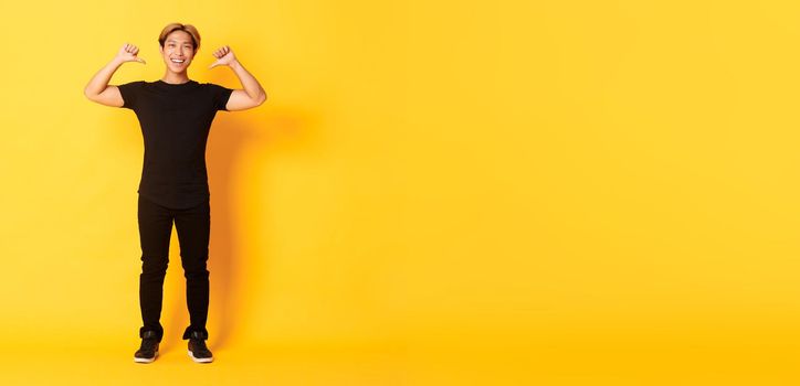 Confident attractive blond asian guy, smiling sassy and pointing at himself, standing over yellow background.