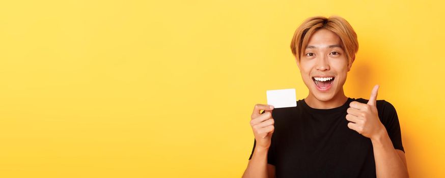 Close-up of happy and satisfied asian handsome guy, showing credit card and thumbs-up in approval, smiling amazed, standing yellow background.