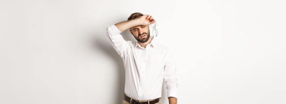 Tired office worker take-off glasses, wiping sweat off forehead with his arm, standing drained against white background.