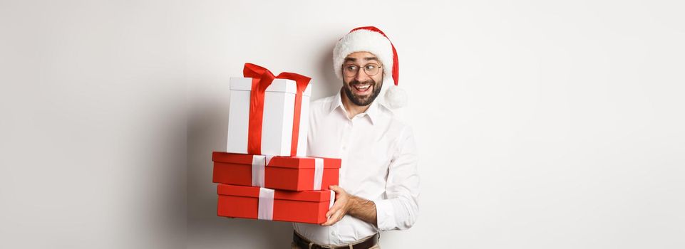 Merry christmas, holidays concept. Excited man celebrating xmas, wearing santa hat and holding gifts, standing over white background.