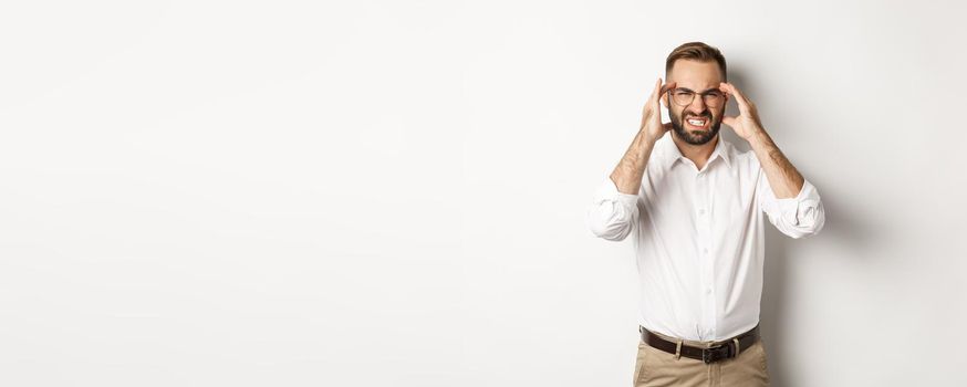 Businessman having headache, grimacing and holding hands on head, standing over white background.