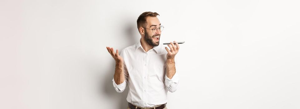 Excited businessman talking on speakerphone and smiling, record voice message with ecstatic face, standing over white background.