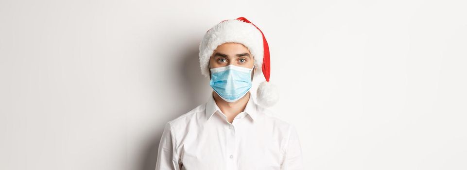 Concept of covid-19, social distancing and winter holidays. Close-up of young man wearing santa hat and face mask from coronavirus, celebrating New Year, white background.