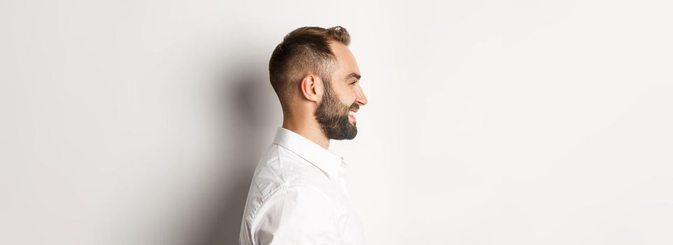 Close-up profile shot of handsome bearded man looking left and smiling, standing against white background.