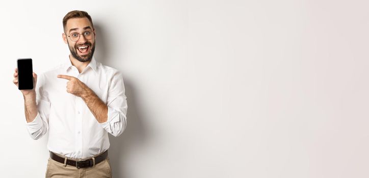 Excited handsome guy showing mobile phone, pointing finger at screen and smiling, standing against white background.