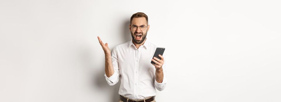 Frusteated man using mobile phone and looking disappointed, complaining, standing over white background.