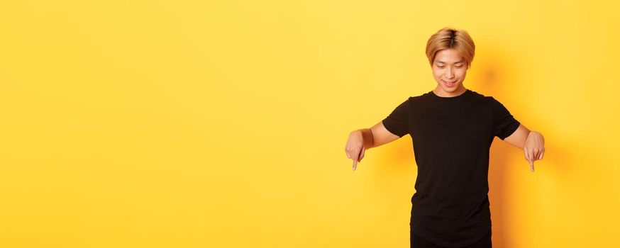 Portrait of curious happy blond asian guy, looking and pointing fingers down with pleased smile, yellow background.