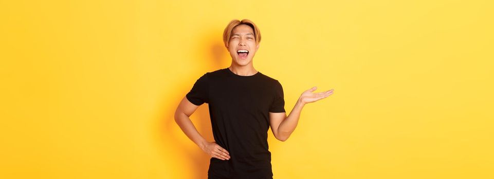 Portrait of happy blond asian guy, winking sassy and smiling, holding something on hand over yellow background.