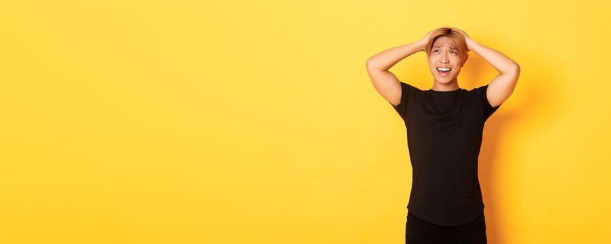 Portrait of young asian guy panicking, looking anxious and upset, holding hands on head and grimacing, standing over yellow background.