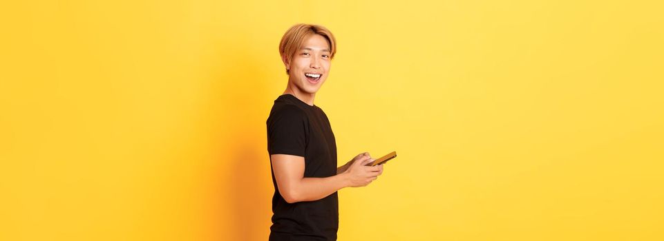 Portrait of handsome stylish asian guy in black outfit, using mobile phone and turning head at camera with satisfied smile, yellow background.