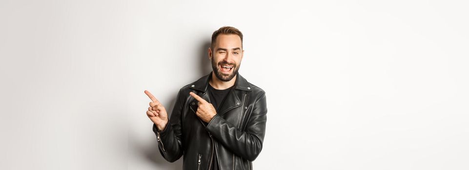 Cheeky guy in leather biker jacket, winking flirty and pointing at upper right corner copy space, white background.