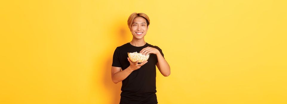 Pleased handsome korean guy smiling happy as enjoying watching movie or tv series, eating popcorn, standing yellow background.