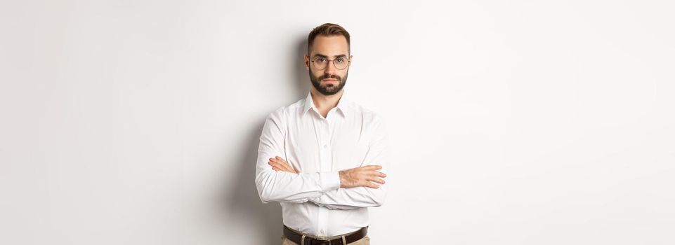 Confident businessman in glasses looking at camera, cross arms on chest, standing over white background.