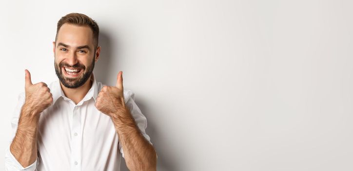 Close-up of satisfied bearded guy in white shirt, showing thumbs up in approval, like and agree, positive answer, white background.