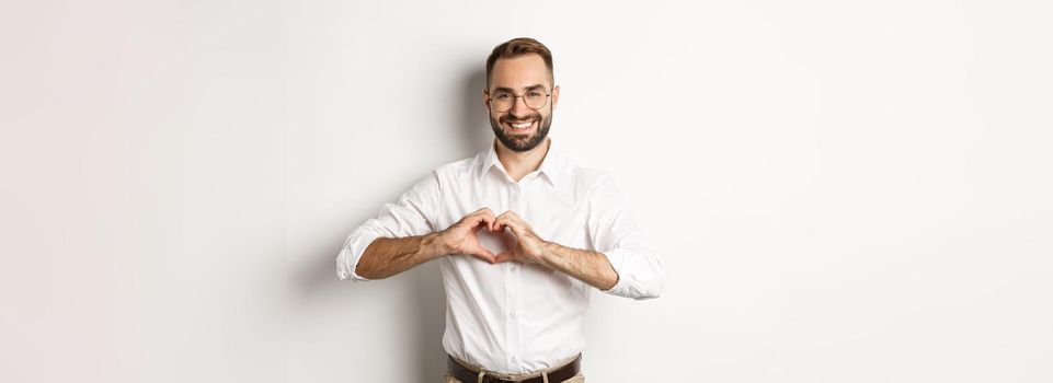 Handsome manage showing heart sign and smiling, I love you gesture, standing over white background.