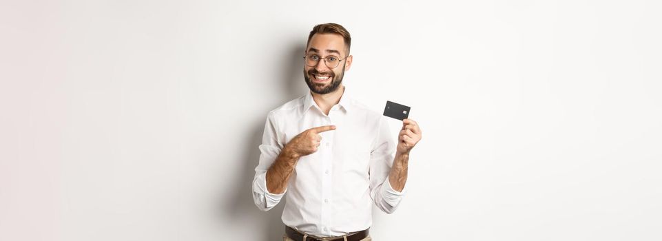 Handsome satisfied man in glasses pointing at credit card, pleased with bank services, standing over white background.