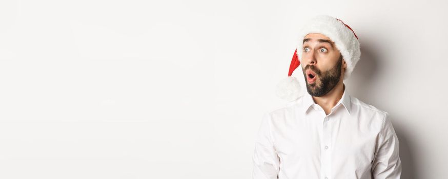 Close-up of amazed bearded man in santa hat, looking left with surprised face, christmas promo offer, standing over white background.