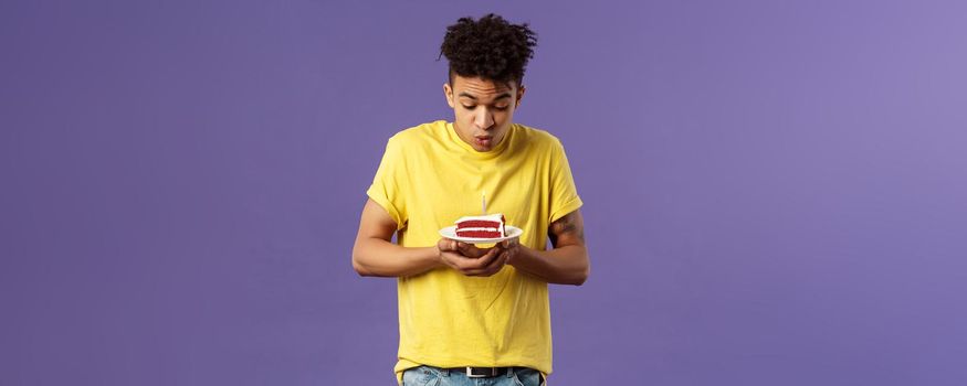 Portrait of cute excited young guy with dreads and tattoos, making wish on birthday, celebrating party, blowing candle on b-day cake, standing purple background dreaming. Holidays concept