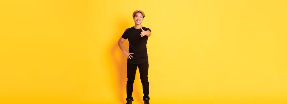 Full-length of friendly handsome korean guy with blond hair, extending hand for handshake, saying hi, standing yellow background.