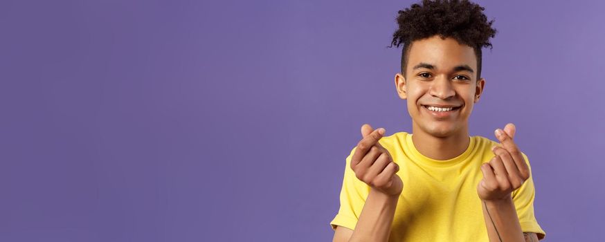 Close-up portrait of lovely cute young man with afro haircut, showing korean heart finger signs and smiling, express his love and sympathy towards person, stand purple background.