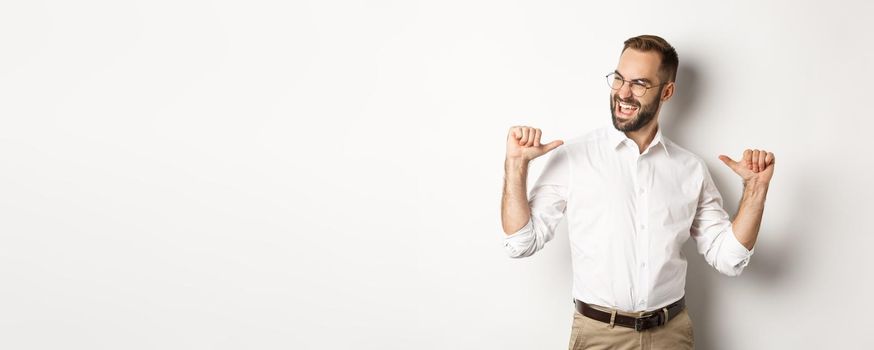 Satisfied and self-assured businessman pointing at himself, standing over white background.