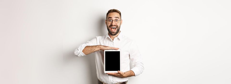 Excited man showing digital tablet screen, smiling amazed, standing over white background.