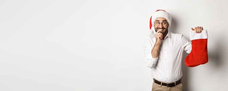 Merry christmas, holidays concept. Adult man looking happy and curious at xmas sock, receive gifts, wearing santa hat, white background.