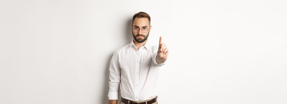 Serious man in glasses showing stop sign, shaking finger to prohibit and forbid, standing over white background.