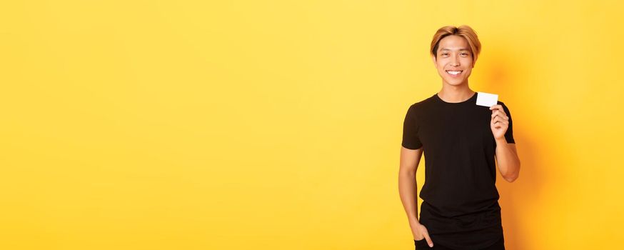 Cheerful handsome asian guy smiling pleased and showing credit card, standing yellow background.