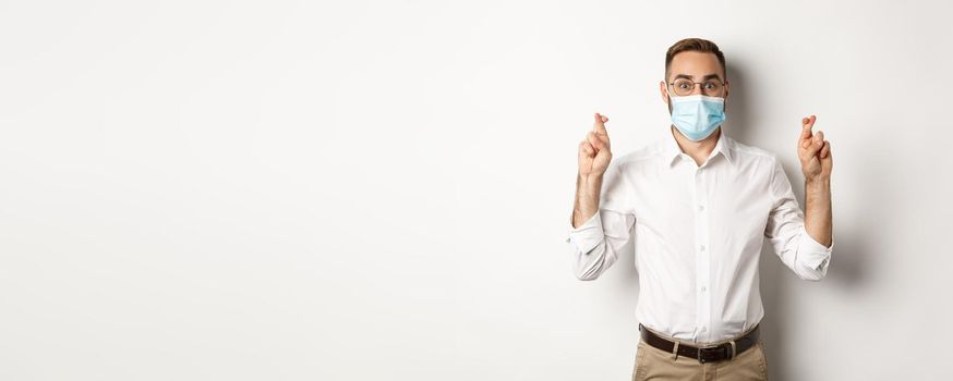 Covid-19, social distancing and quarantine concept. Hopeful employee in medical mask, holding fingers crossed and making wish, white background.