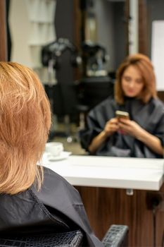 Beautiful young red hair woman using her smartphone and texting sitting in front of a mirror waiting to visit a hairdresser