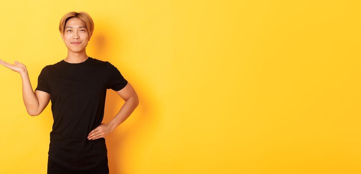 Portrait of happy and proud smiling asian guy holding something on hand over yellow background.