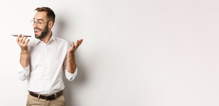 Excited businessman talking on speakerphone and smiling, record voice message with ecstatic face, standing over white background.