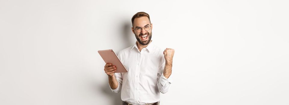 Successful businessman rejoicing on winning online, reading on digital tablet and making fist pump, triumphing, standing over white background.