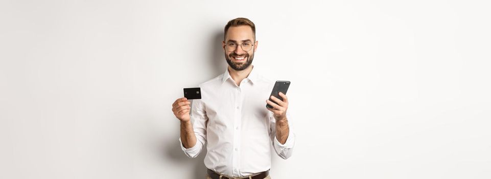 Business and online payment. Smiling male entrepreneur shopping with credit card and mobile phone, standing over white background.