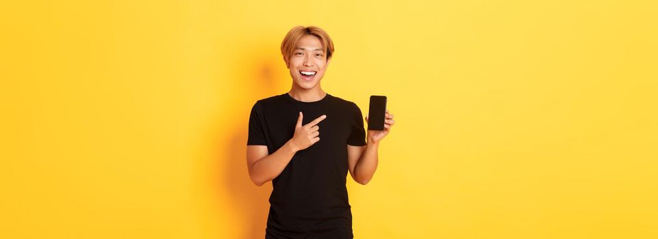 Portrait of satisfied smiling asian guy with blond hair, looking amused and pointing finger at smartphone screen, standing yellow background.