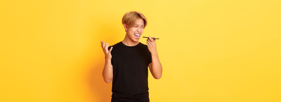 Portrait of angry asian man yelling mad at mobile phone speaker, record furious voice message, standing yellow background.