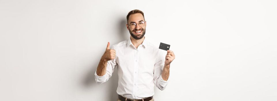 Handsome man showing his credit card and thumb up, recommending bank, standing over white background. Copy space