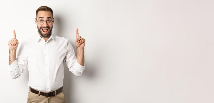 Excited businessman checking out advertisement, pointing and looking up with happy face, standing against white background.