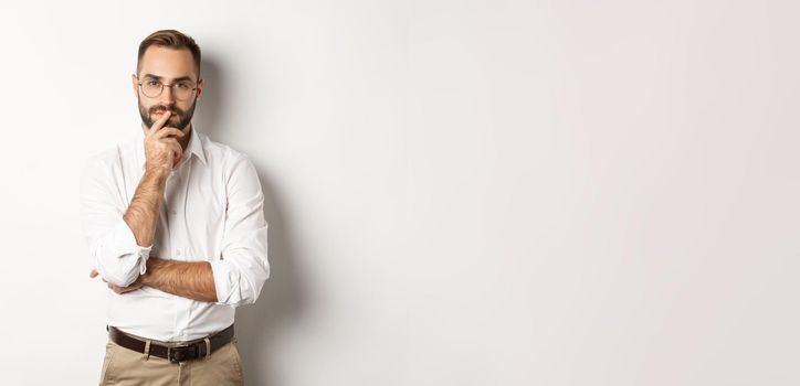 Thoughtful handsome businessman looking at camera, making choice or thinking, standing in glasses and white collar shirt against studio background.