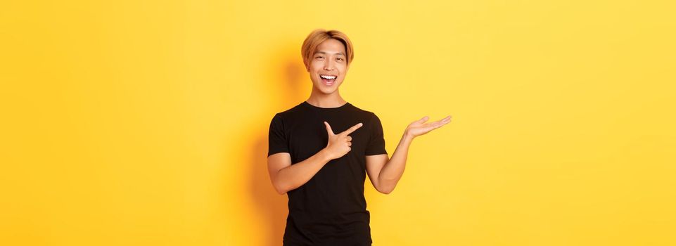 Portrait of handsome smiling asian guy holding something on hand and pointing finger at it, standing yellow background pleased.