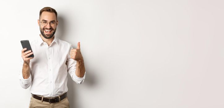 Satisfied business man showing thumbs up after using mobile phone, standing pleased over white background.