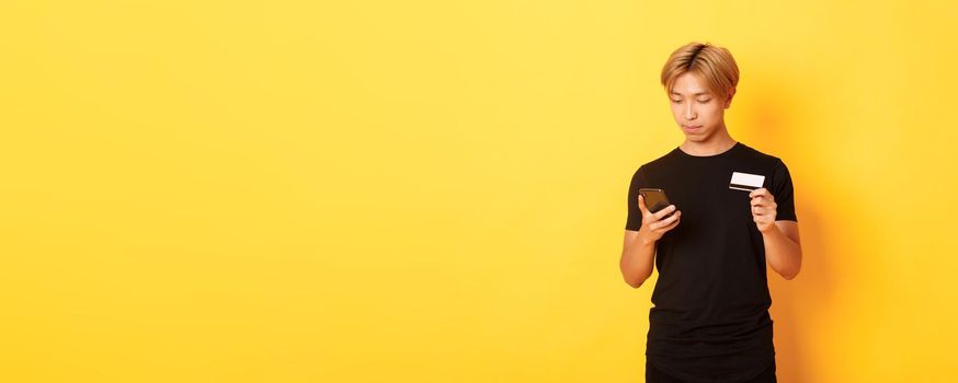 Young handsome asian guy in black t-shirt shopping online, holding credit card and using smartphone, standing yellow background.