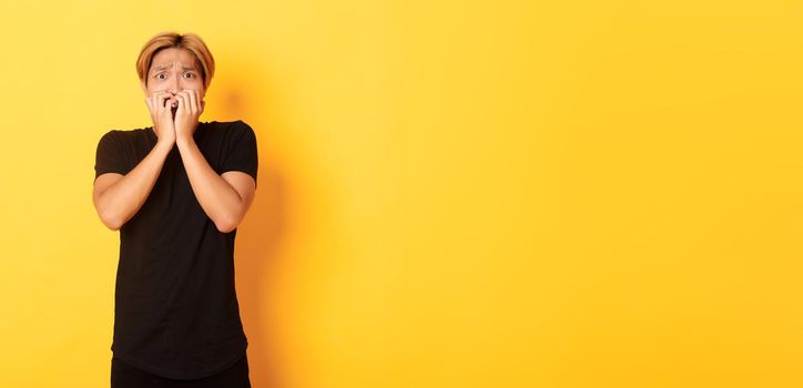 Portrait of scared insecure asian blond guy, holding hands over mouth horrified, looking frightened, yellow background.