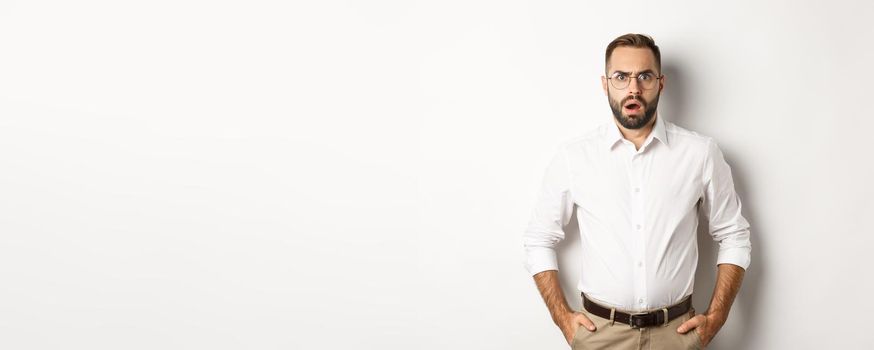 Shocked and displeased businessman in glasses, gasping and looking upset at camera, standing over white background. Copy space