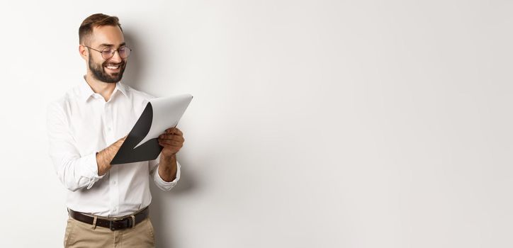 Man looking satisfied while reading documents, holding clipboard and smiling, standing over white background.