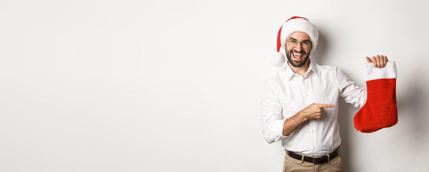 Happy man in santa hat celebrating winter holidays, pointing at christmas sock and smiling, holding gifts, standing over white background.