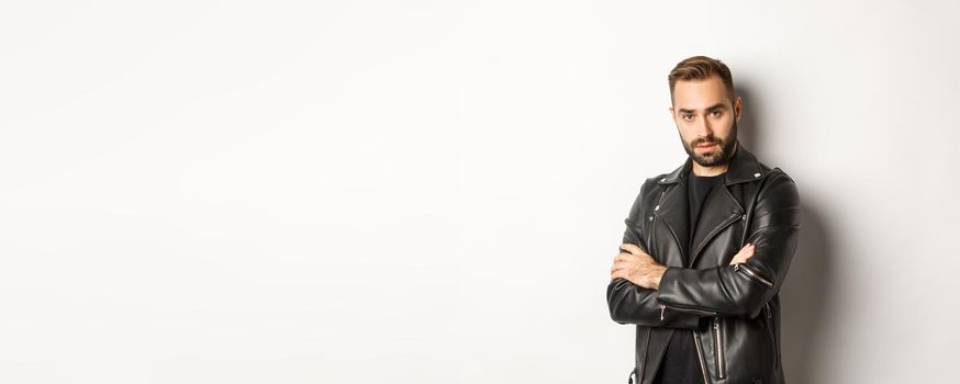 Attractive macho man with beard, wearing leather jacket smiling, looking confident with hands crossed on chest, white background.