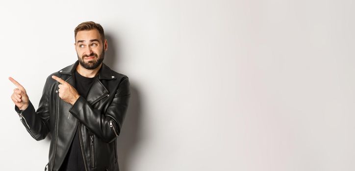 Skeptical and doubtful guy in black leather jacket, shrugging while pointing at upper left corner promo offer, standing over white background.